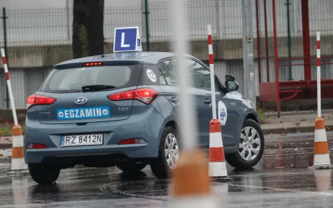 Samochód egzaminacyjny z literą "L" na dachu i napisem "EGZAMIN" na tylnej klapie, otoczony pachołkami na mokrym placu.