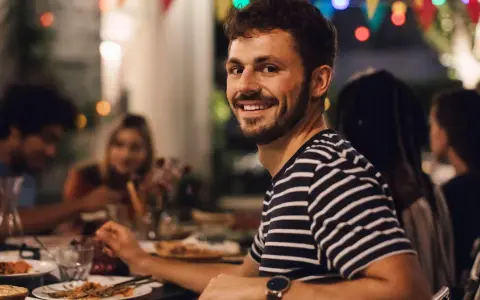 Uśmiechnięty mężczyzna na tle przyjaciół przy stole. Idealny, tani pomysł na wieczór kawalerski z jedzeniem i dobrym towarzystwem.