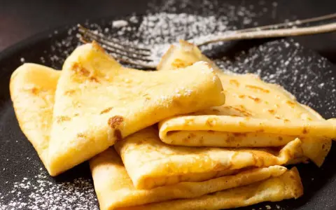Deliciosos crepe en francés, doblados y espolvoreados con azúcar glas sobre un plato negro.