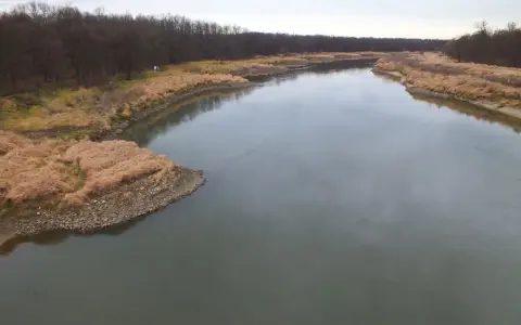 Szeroka rzeka z ciemną wodą, otoczona brzegami porośniętymi suchą trawą i lasem. Widać głębokie miejsce w rzece.