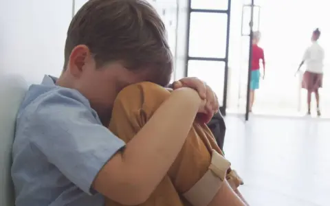 Smutny chłopiec skulony na podłodze. Nie chce chodzić do szkoły, czuje się samotny, podczas gdy inni bawią się w tle.
