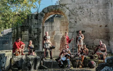 Un groupe de guerriers, prêts à combattre, pose devant des ruines antiques. Ces hommes, tels des gladiateurs romains, arborent armures et armes, prêts pour l'arène.