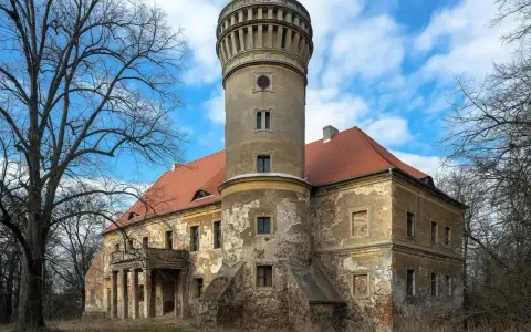 Zniszczony pałac w Osetnie z charakterystyczną wieżą i czerwoną dachówką, otoczony drzewami.