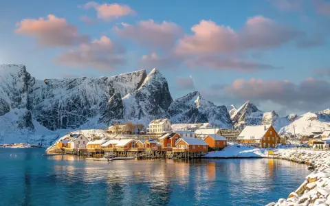 Snow-covered mountains frame a picturesque village with orange and white houses on stilts over the water, a scene from one of Europe's best winter destinations.