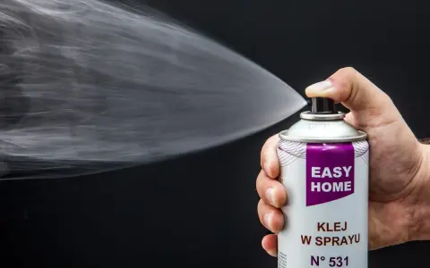 Dłoń trzyma puszkę z klejem w sprayu, który jest rozpylany. Idealny klej do podsufitki bez zdejmowania.