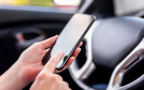 Ręce trzymają smartfon w samochodzie, obok kierownicy. Może to być pomocne, by dowiedzieć się, co to aux i jak podłączyć muzykę.