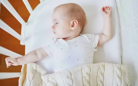 Baby schläft im Gitterbett, das Baby bewegt sich viel im Schlaf und streckt die Arme aus.