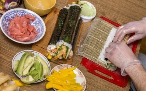 Ręce w rękawiczkach zwijają sushi na macie bambusowej. Obok gotowe rolki, awokado, mango i marynowany imbir. Oto jak się robi sushi!