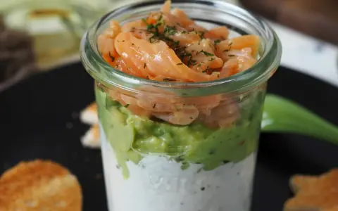 Leckerer Lachs Avocado Vorspeise im Glas, geschichtet mit cremiger Sauce und garniert mit Dill. Dazu geröstete Brotscheiben.