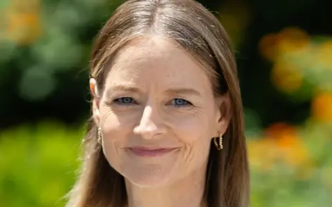 Porträt einer Frau mit blauen Augen und braunem Haar, das einige einzelne graue Haare abdeckt. Sie trägt goldene Ohrringe.