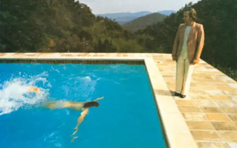A portrait of an artist by a pool with two figures. One figure swims in the bright blue water, while another stands on the tiled edge, looking out at the mountainous landscape.