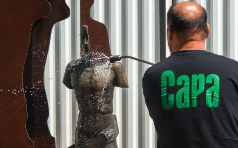 Hombre con camiseta negra con letras verdes "Capa" limpia una estatua de bronce con una manguera a presión.
