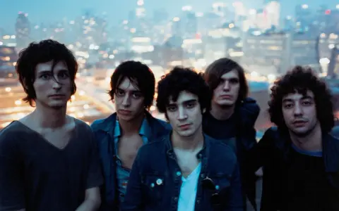 Les membres de The Strokes posent ensemble, le regard tourné vers l'objectif, avec une ville illuminée en arrière-plan.