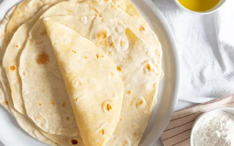 Piatto di piadine senza strutto, morbide e dorate, pronte per essere farcite. Accanto, una ciotolina d'olio e una di farina.