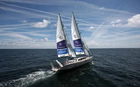 Le voilier Pangaea de Mike Horn navigue sur une mer calme sous un ciel bleu parsemé de nuages et de traînées d'avions.
