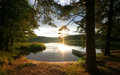 Zachód słońca nad jeziorem na Mazurach. Piaszczysta plaża, łódka przy brzegu, drzewa i odbijające się w wodzie światło.