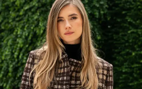 Mujer con cabello rubio y mechas invertidas, viste un abrigo de cuadros marrones y un cuello alto negro. Fondo verde frondoso.