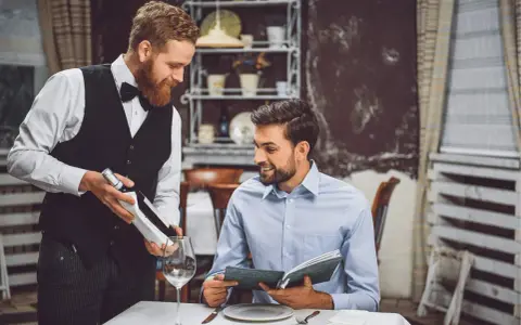 Camarero atento presenta una botella de vino a un cliente que revisa el menú. Demuestra las cualidades de un camarero: servicio, amabilidad y conocimiento.