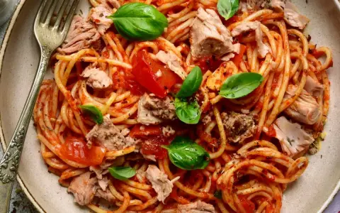 Spaghetti al sugo di pomodoro con abbondante tonno in scatola, guarniti con foglie di basilico fresco. Un piatto semplice e gustoso.