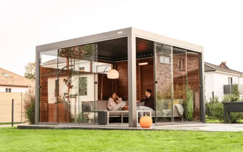 Un couple se détend dans une pergola moderne avec parois vitrées. La fixation pergola est discrète, laissant place à un espace cosy avec chauffage d'appoint.