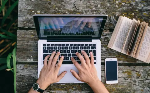 Ręce piszą CV funkcjonalne na laptopie, obok leży otwarta książka i telefon.