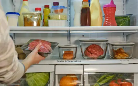 Mano colocando carne picada envasada en un refrigerador lleno de alimentos.
