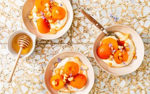 Trois bols de compote abricot onctueuse, garnie d'amandes et de fleurs violettes. Un pot de miel avec sa cuillère est à côté.