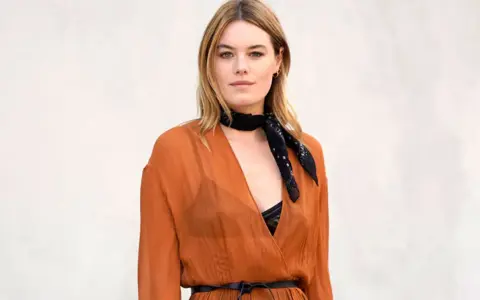 Camille Rowe luce un pañuelo negro y un vestido naranja con escote pronunciado.