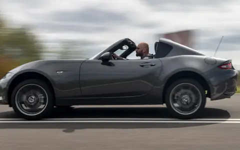 Mężczyzna w szarej kurtce prowadzi ciemnoszarą Mazdę Miata z opuszczonym dachem, jadąc po drodze.