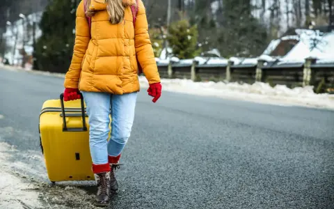 Kobieta w żółtej kurtce i czerwonych rękawiczkach ciągnie żółtą walizkę. W tle zimowy krajobraz. Modlitwa o szczęśliwą podróż.