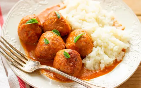 Albondigas a la jardinera con arroz blanco, decoradas con hojas verdes. Un tenedor plateado descansa sobre el plato.