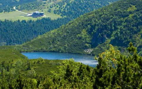 Widok na górskie jezioro i schronisko Samotnia w Karkonoszach. Otoczone zielenią, lasem i skalistymi zboczami.