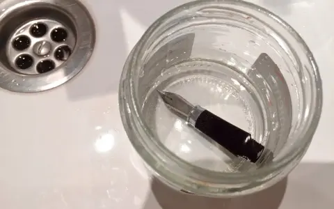 Pluma estilográfica sumergida en un frasco de vidrio con agua, junto a un desagüe. Un paso para aprender como limpiar una pluma.