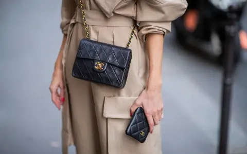Elegancka kobieta w beżowym płaszczu prezentuje klasyczną, pikowaną torebkę Chanel vintage z złotym łańcuszkiem i logo.