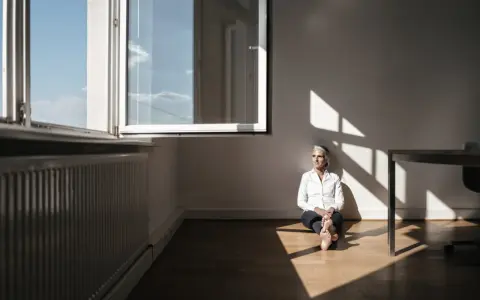 Kobieta w białej koszuli siedzi na podłodze w pustym pokoju, ciesząc się światłem wpadającym przez okno. To kwintesencja minimalizmu w domu.
