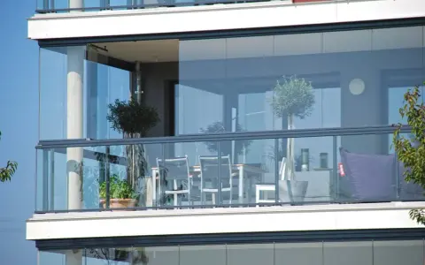 Un balcone moderno con vetrate scorrevoli, ideale per chiudere un balcone fai da te. Tavolo, sedie e un pouf viola creano un angolo relax.