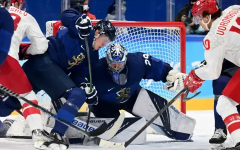 Bramkarz Finlandii broni strzał. W tle zawodnicy walczą o krążek, dążąc do zwycięstwa, jak przystało na mistrzów olimpijskich w hokeju na lodzie.