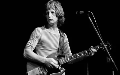 Pete Ham, mit gestreiftem Shirt und Gitarre, blickt konzentriert auf die Bühne.