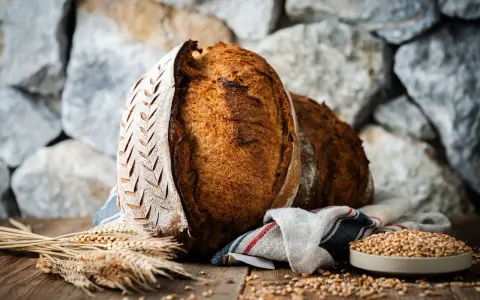 Zwei rustikale Brote, eines mit eingekerbtem Muster, liegen auf einem Holztisch. Weizenähren und eine Schale voller Körner deuten auf ein köstliches Ruchbrot Rezept hin.
