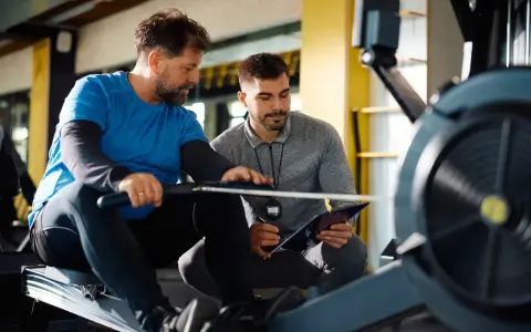 Trener personalny omawia z klientem postępy na tablecie, siedząc na wioślarzu. Zastanawiasz się, ile zarabia trener personalny?