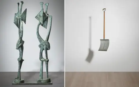 Two abstract bronze figures stand side-by-side, a modern sculpture. To the right, a metal snow shovel hangs suspended, its shadow cast on the wall.