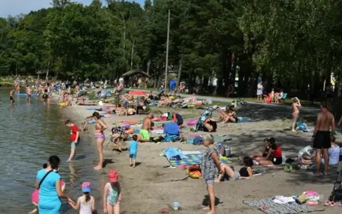 Tłoczna plaża nad jeziorem na Pomorzu. Ludzie opalają się, kąpią i odpoczywają w cieniu drzew.