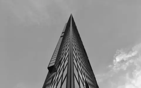 Minimalistyczne fotografowanie architektury. Ostry szczyt wieżowca przecina niebo, odbijając chmury w szklanej fasadzie.