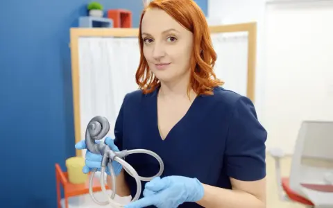 Kobieta w niebieskim fartuchu trzyma model ucha, pokazując jego budowę, w tym ślimak, który przypomina kształtem nerw słuchowy.