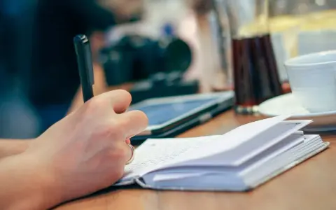 Mano escribiendo en un cuaderno abierto, inspirada para escribir cuentos. En la mesa hay una tableta, una taza y una jarra.