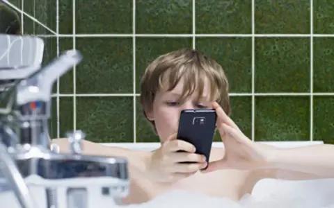 Chłopiec w wannie z telefonem. To jeden z objawów uzależnienia od telefonu u dzieci, gdy nawet kąpiel nie przerywa kontaktu z ekranem.