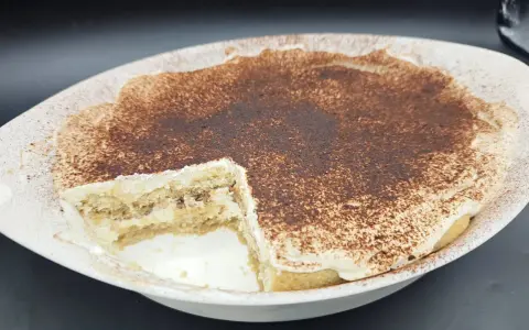Un délicieux tiramisu sans oeuf, crémeux et saupoudré de cacao, dans un plat blanc. Une part a été retirée, révélant les couches de biscuits et de crème.