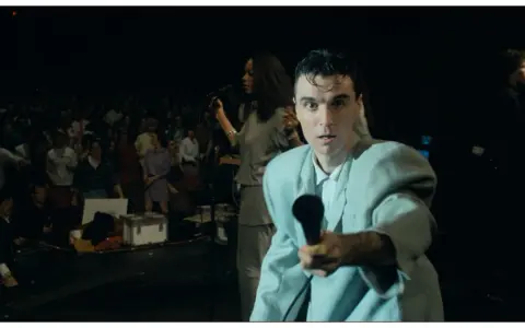 David Byrne z The Cult - Be Free, śpiewa do mikrofonu na tle tłumu.