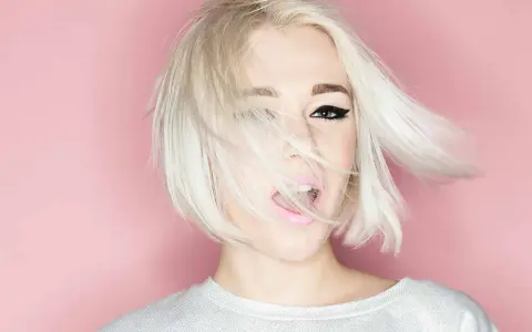 Mujer con cabello rubio platino y corte recto al hombro, con un ojo visible y labios rosados abiertos.
