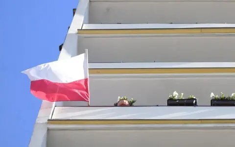 Prawidłowa flaga Polski powiewa na tle błękitnego nieba, obok balkonów z kwiatami.